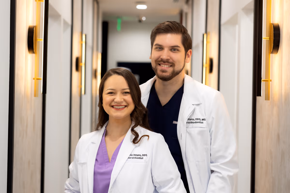Smiling female and male dentists in white coats standing in a hallway with wall lights.