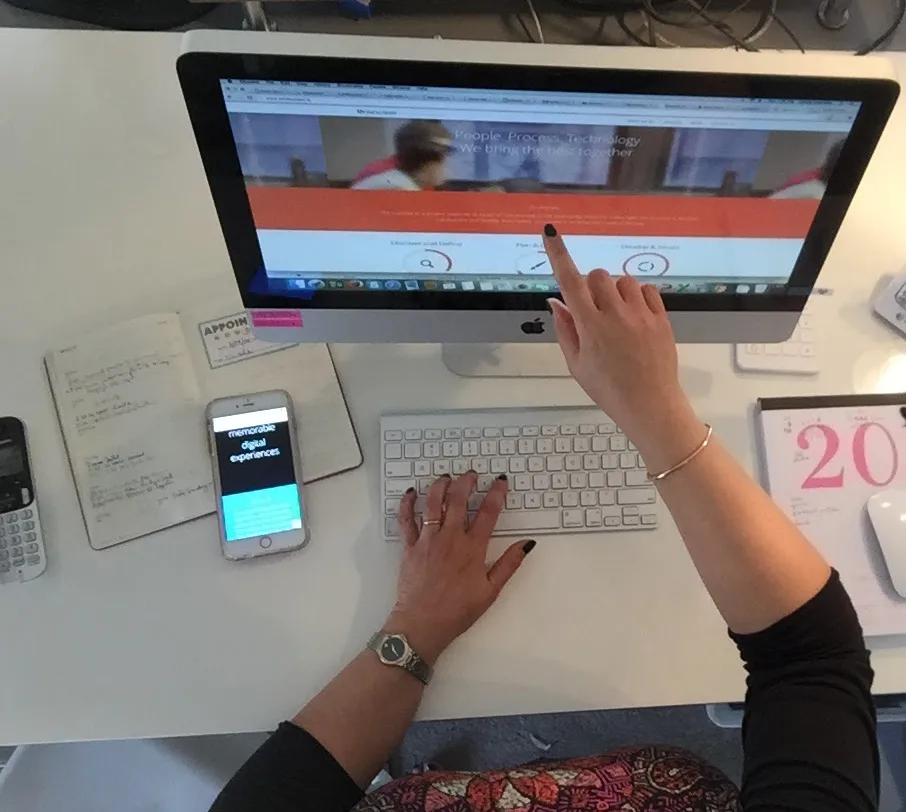 Overhead view of a woman reviewing a professionally designed website on her iMac, with the same site open on her phone beside a notebook