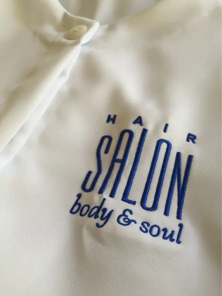 Professional white esthetician gown with custom blue embroidered salon logo for personal care uniforms