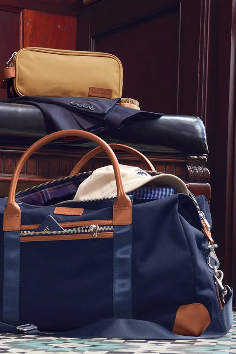 Brooks Brothers navy canvas duffel bag with tan leather trim, packed with a cap, dress shirt, and travel kit