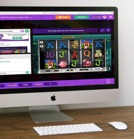 Desktop computer displaying an online casino slot game interface with colorful reels, chat panel, and rewards navigation, set on a desk with keyboard and mouse.