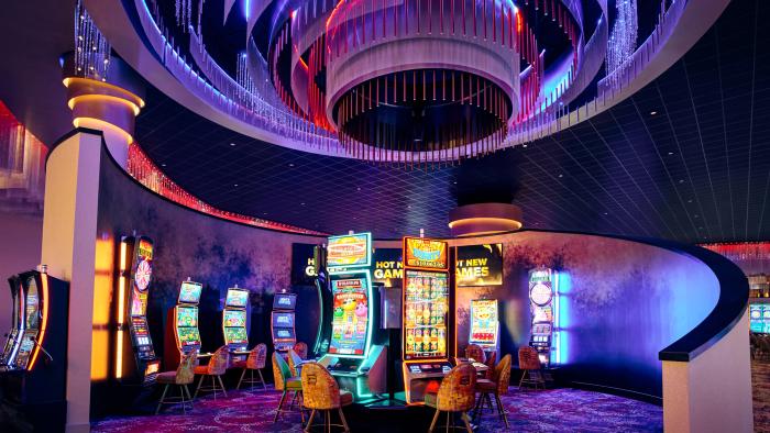 Interior of a casino with colorful slot machines, patterned carpet, and a large illuminated chandelier overhead.
