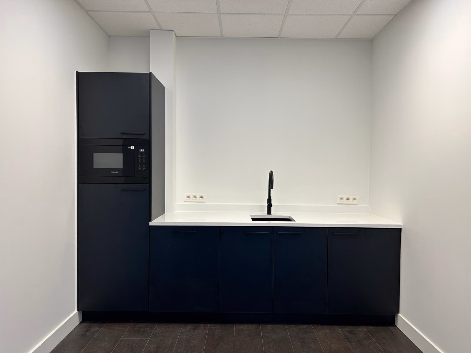 Zwarte kitchenette met inbouwmicrogolf en wit werkblad in een kantoorruimte