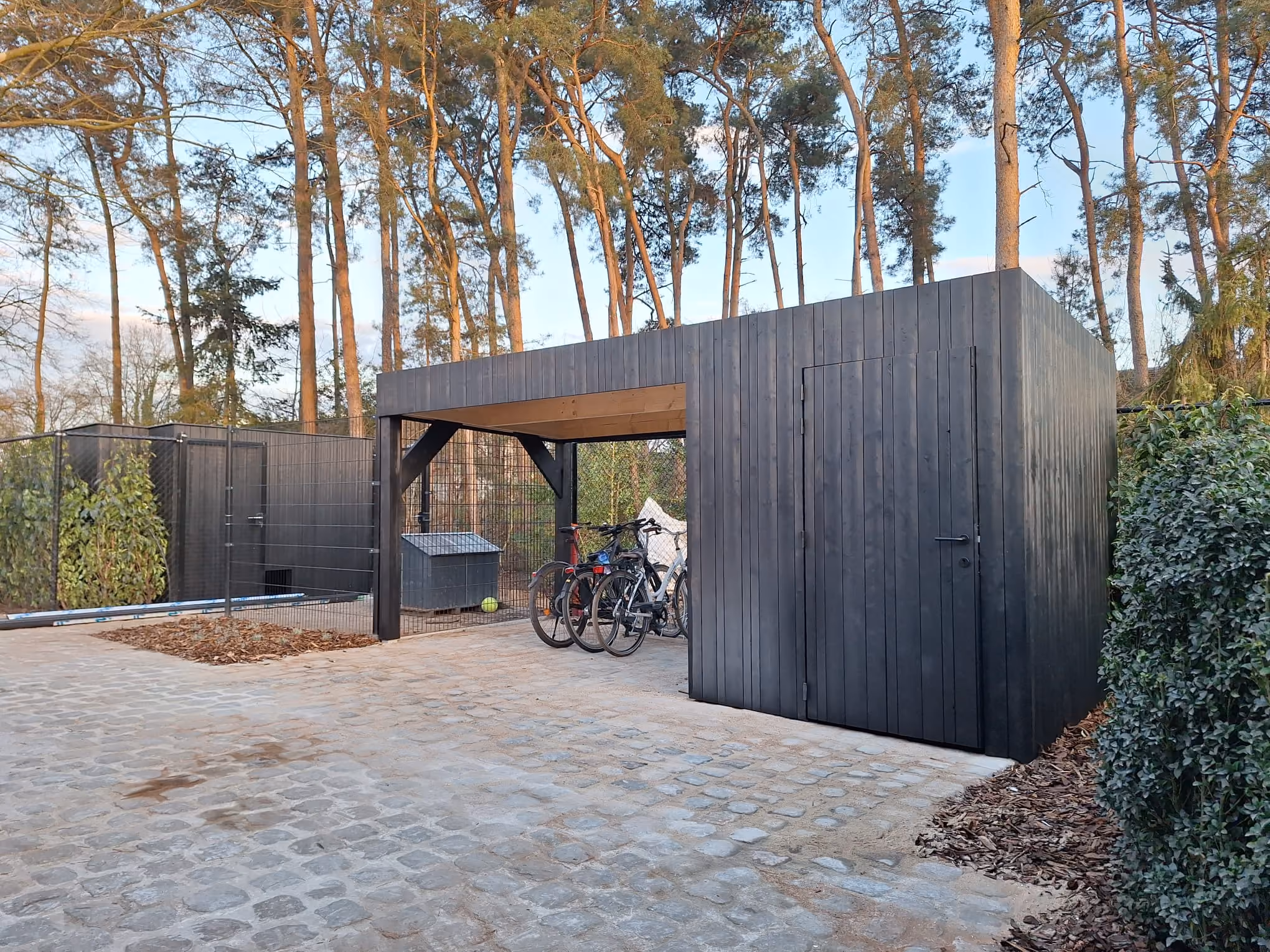 Houten carport en berging met zwarte gevelbekleding tussen de dennenbomen