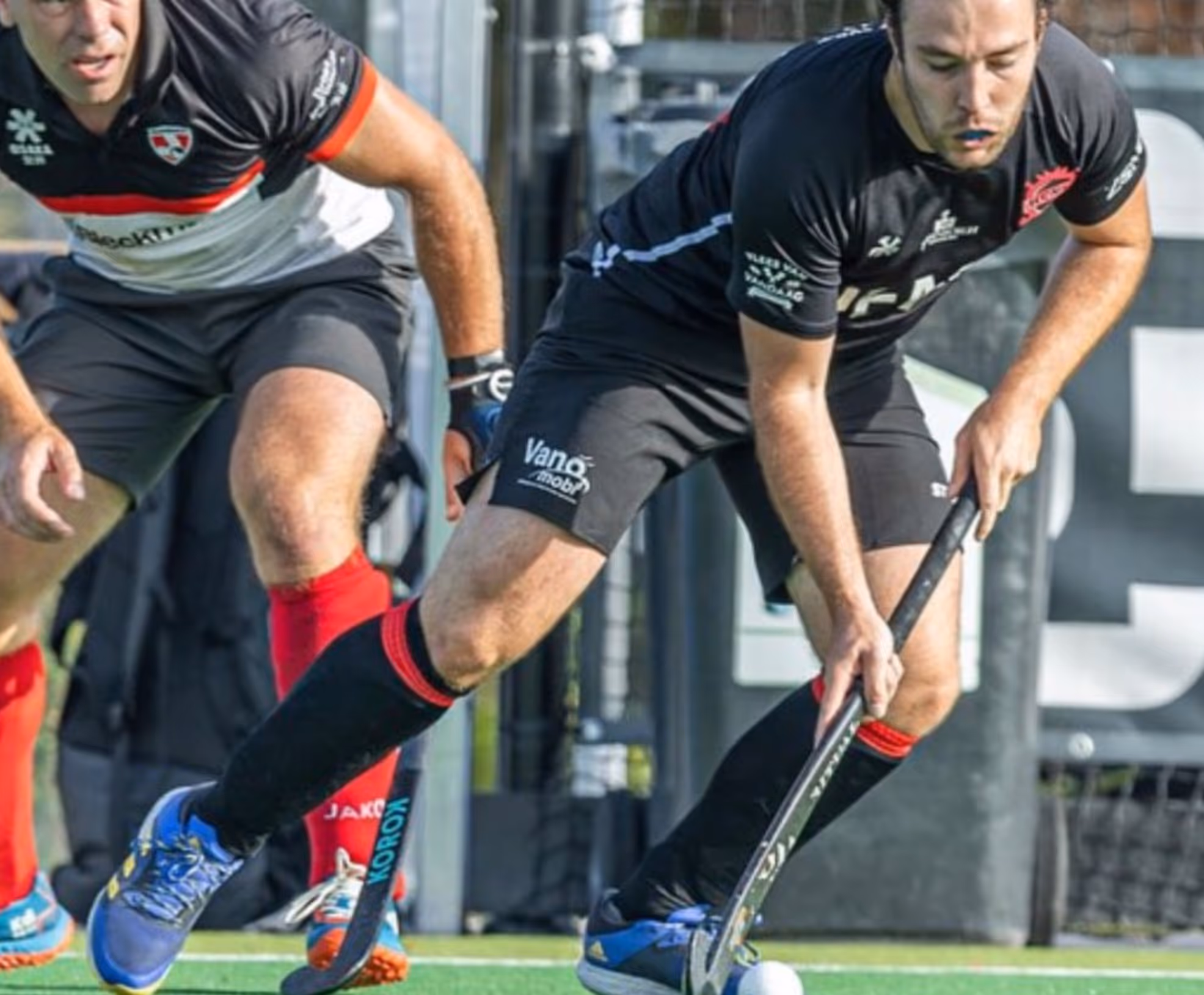 Twee hockeyers, gekleed in zwarte en rode uniformen, zijn in actie op het veld. De ene is gefocust op het controleren van de bal met zijn stick.