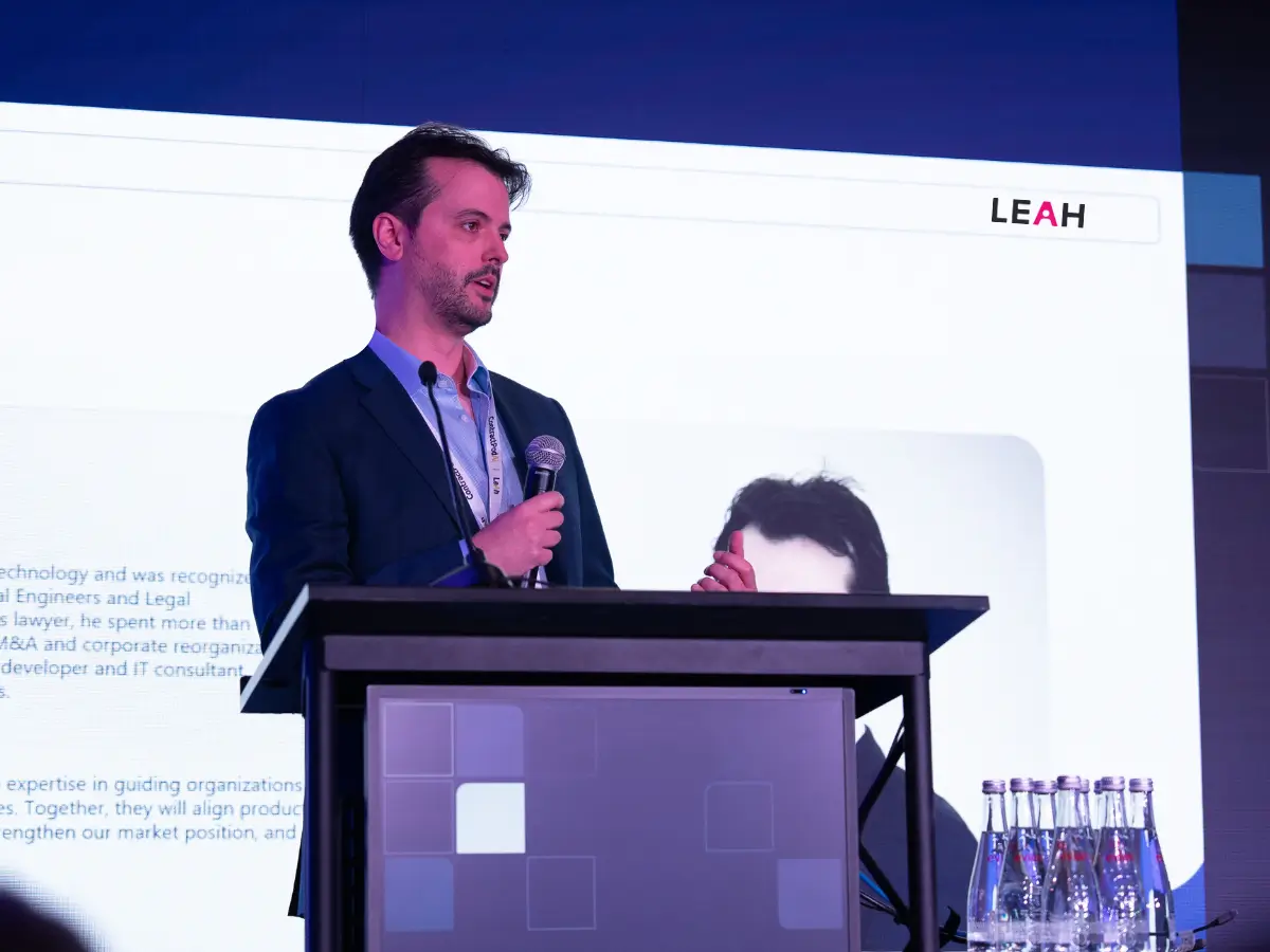 James Thomas, Chief AI Officer at LEAH, speaks at a podium with a microphone during a tech event, with a LEAH-branded slide behind him.