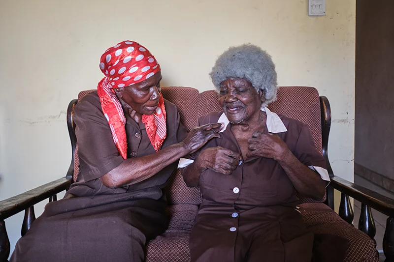 ekesayi Idah Hwiza, a 90-year-old former liberation fighter from Zimbabwe’s independence struggle, is assisted by her longtime friend, Shelta Nhengo, as she fastens the buttons of her uniform inside her house in Mbare township. 