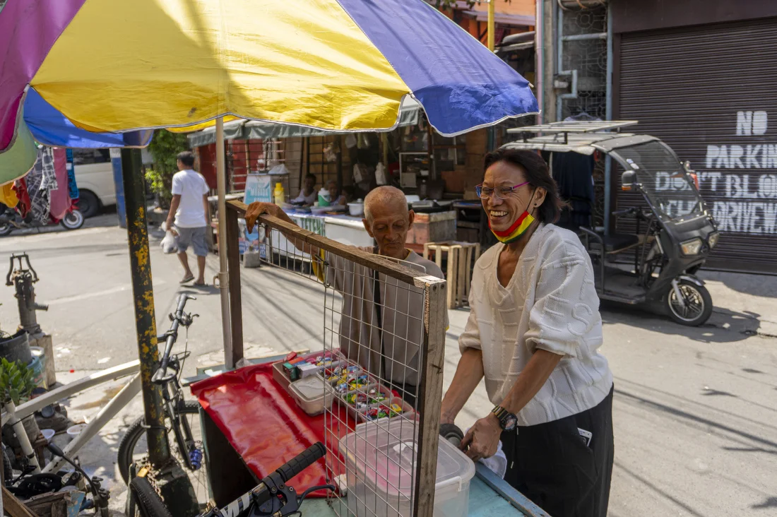 Rey Ravago (64) visits his friend and fellow drag queen, Al Enriquez (88), at his snack and cigarette cart outside his home in Baclaran. Metro Manila, Philippines. June 9, 2024.