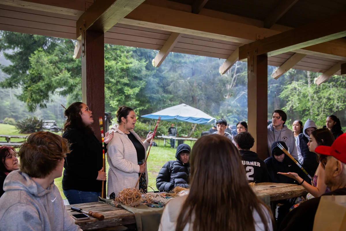 Youth listen as regalia and cultural teachings are shared beneath the shelter.