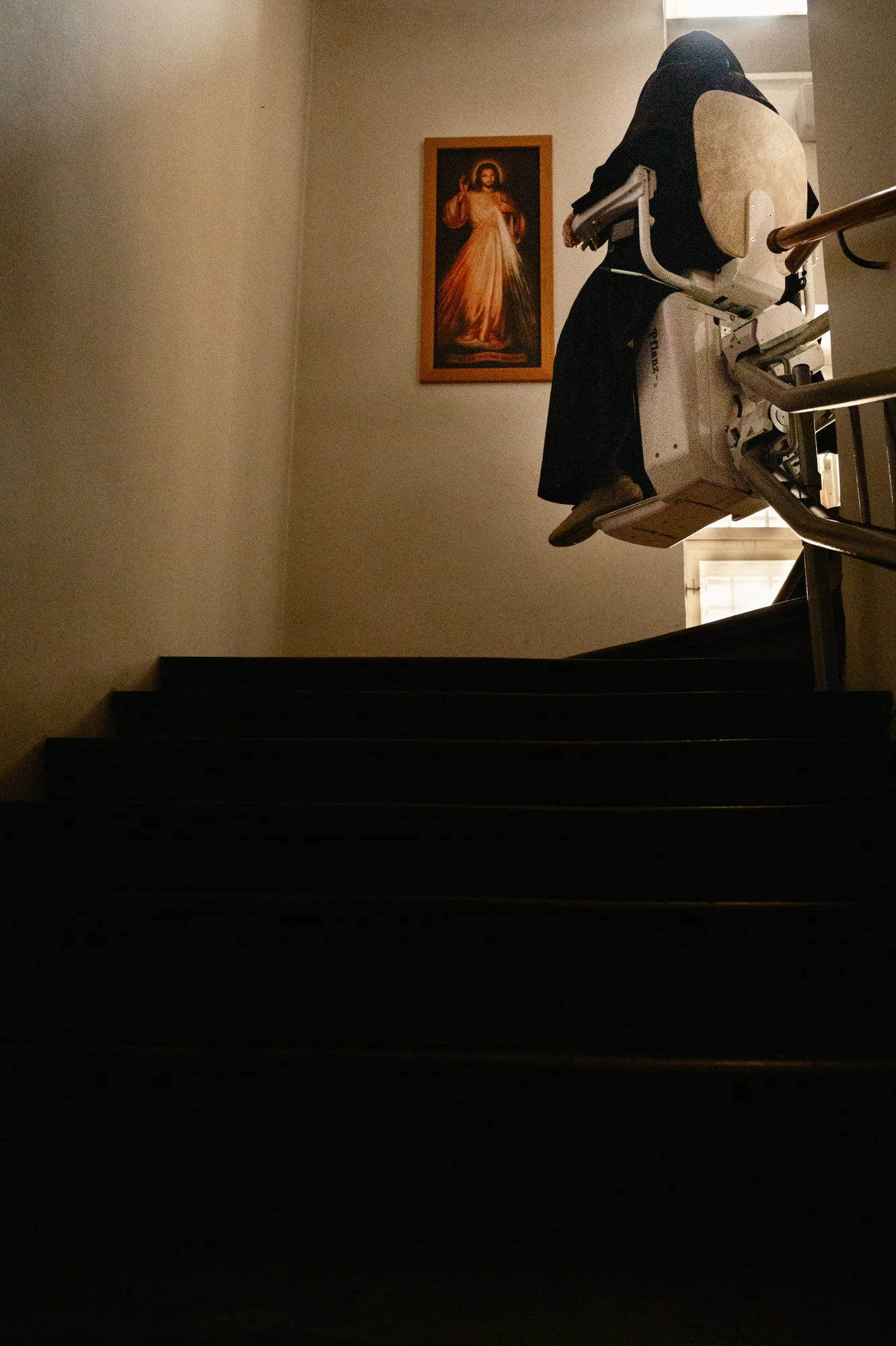 Sister Bernadette, secured in a stair lift, passes a painting of Christ as she ascends up the convent's many floors.