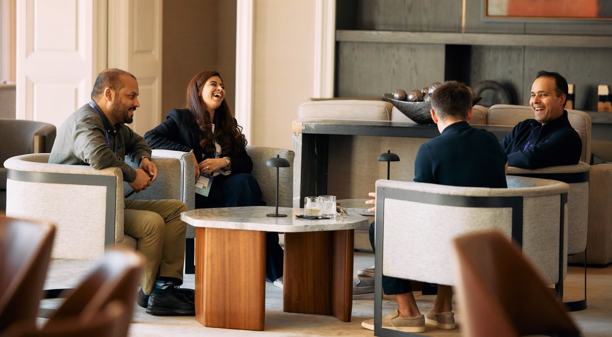Four people sitting in armchairs around a table, smiling and engaging in a lively conversation.