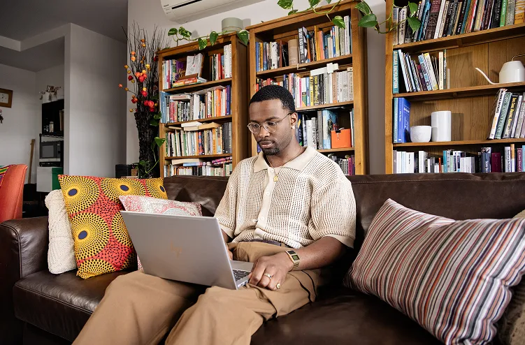 A man looking at a laptop while sitting on the couch with a bookshelf in the background.