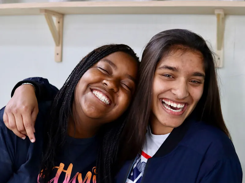 Two girls laugh and smile, one puts her arm over the other