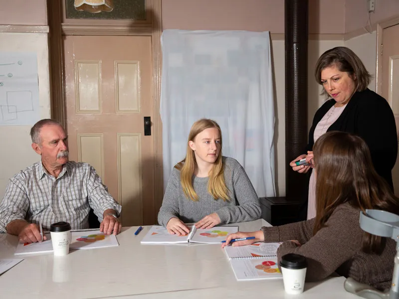 Four people gather around at a table. Three people are sitting, with booklets and pens, while one stands. They all look at each other