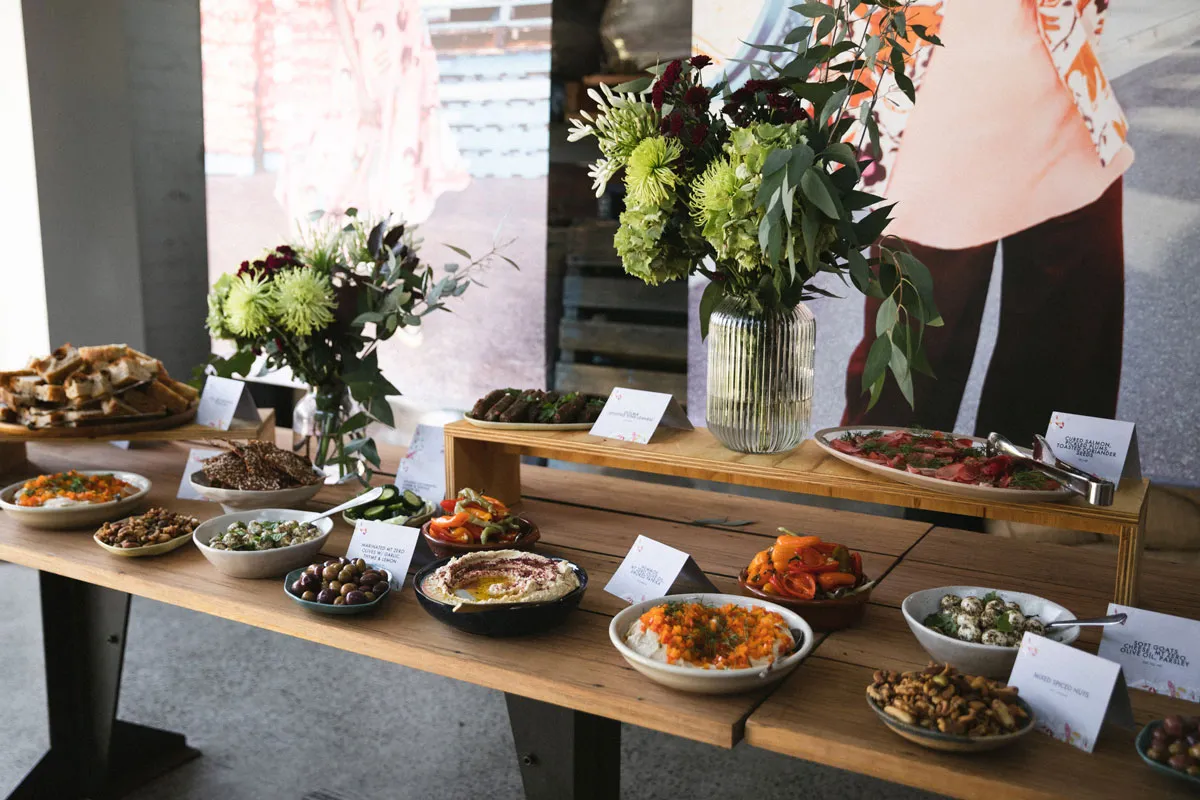 A wooden dinner table is decorated with flowers. The table has a large range of foods from different cultures, with signs about the dish