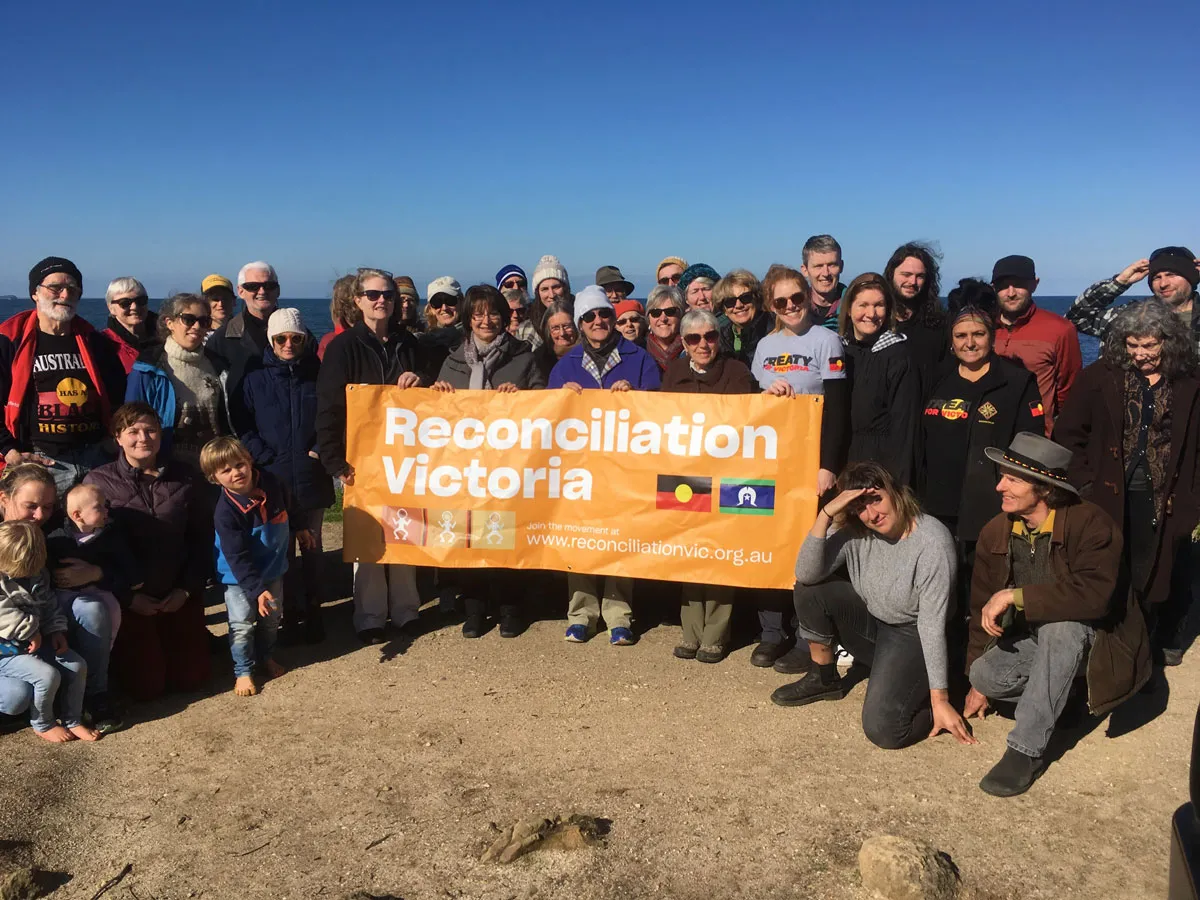 Reconciliation Victoria team stand together at a beach. They hold a sign that reads "Reconciliation Victoria"
