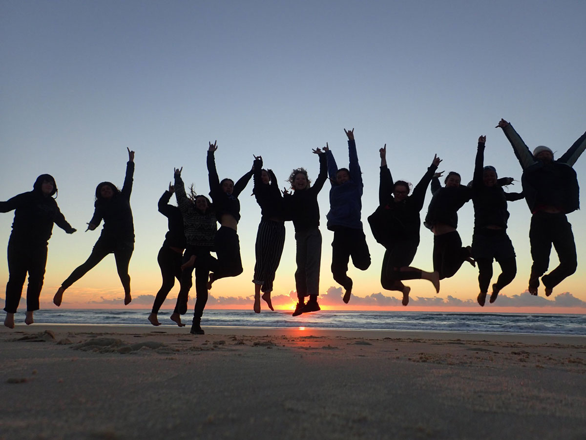 A group of people jump together in the air on the beach at sunset