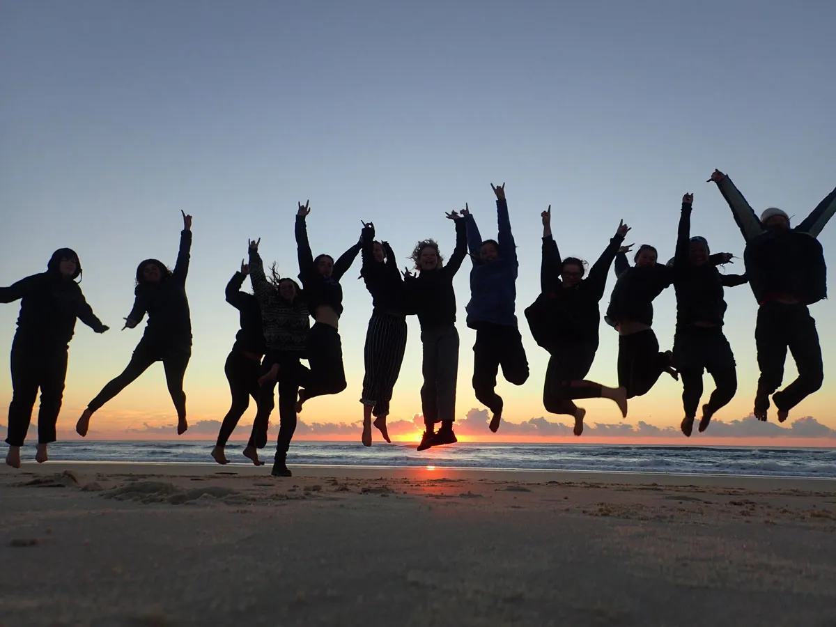 A group of people jump together in the air on the beach at sunset