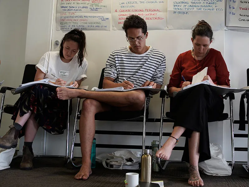 Three people sit on chairs, writing on books