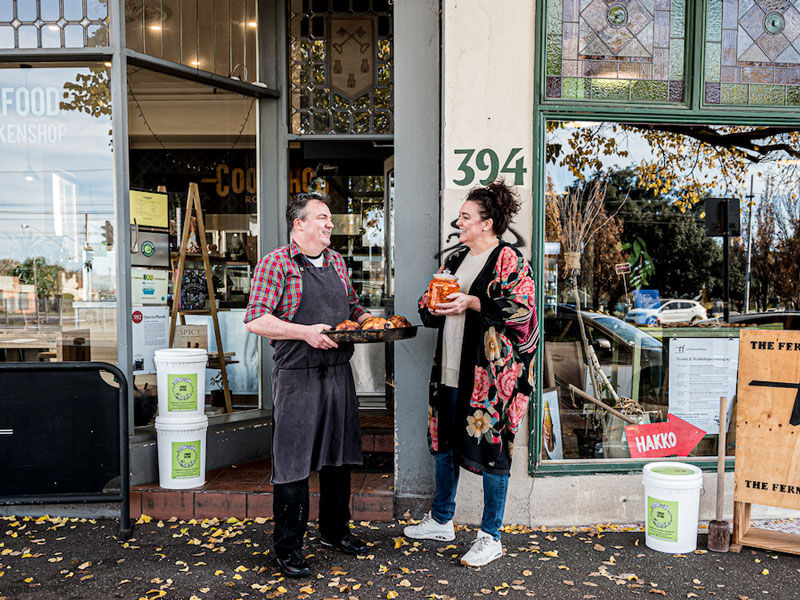 Two people stand outside of a store. One holds a tray of pasties, the other a jar