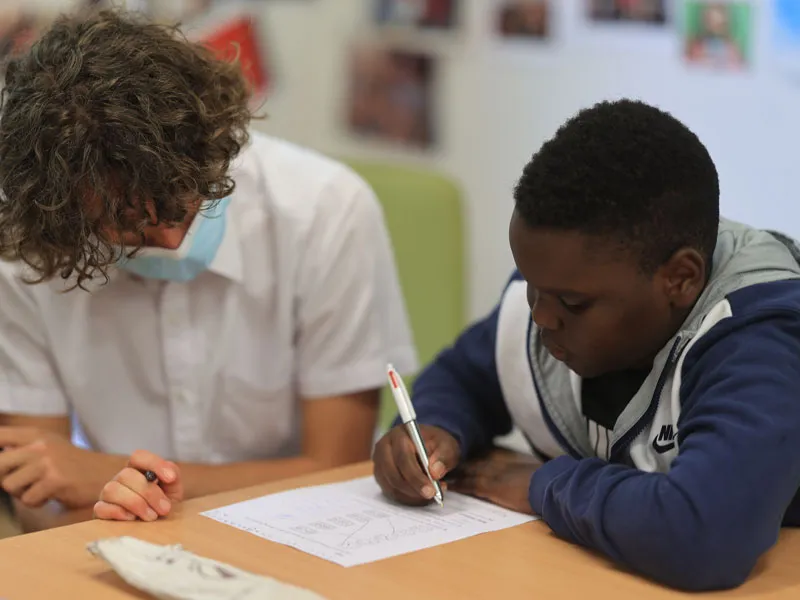 A teacher assists a student in his learning. They both sit at a desk, while the student holds a pen, writing an answer down on a sheet of paper