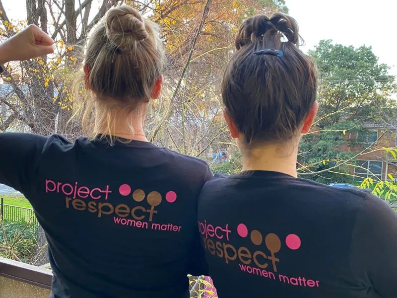 Two woman turn around to show the back of their t-shirts. The shirt says "project respect, women matter". One woman raises her arm in a fist