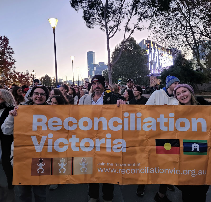 Three people hold up an orange sign during a protest that says "Reconciliation Victoria"