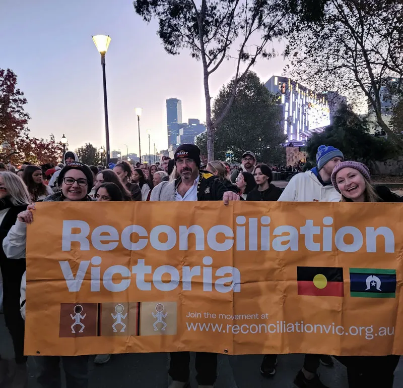 Three people hold up an orange sign during a protest that says "Reconciliation Victoria"