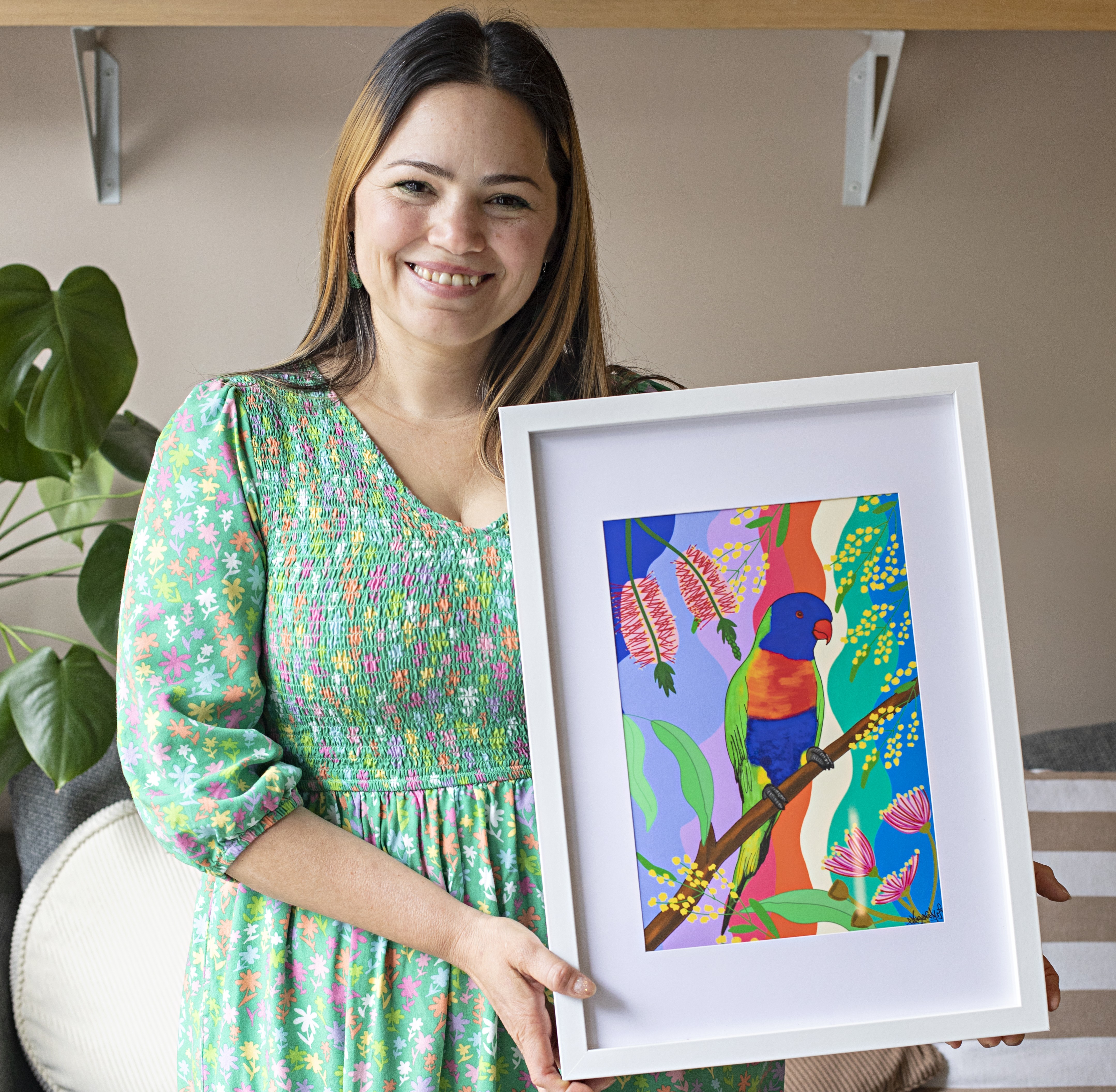 A woman in a green patterned dress holding a framed, colourful painting of a bird surrounded by bright botanical patterns.