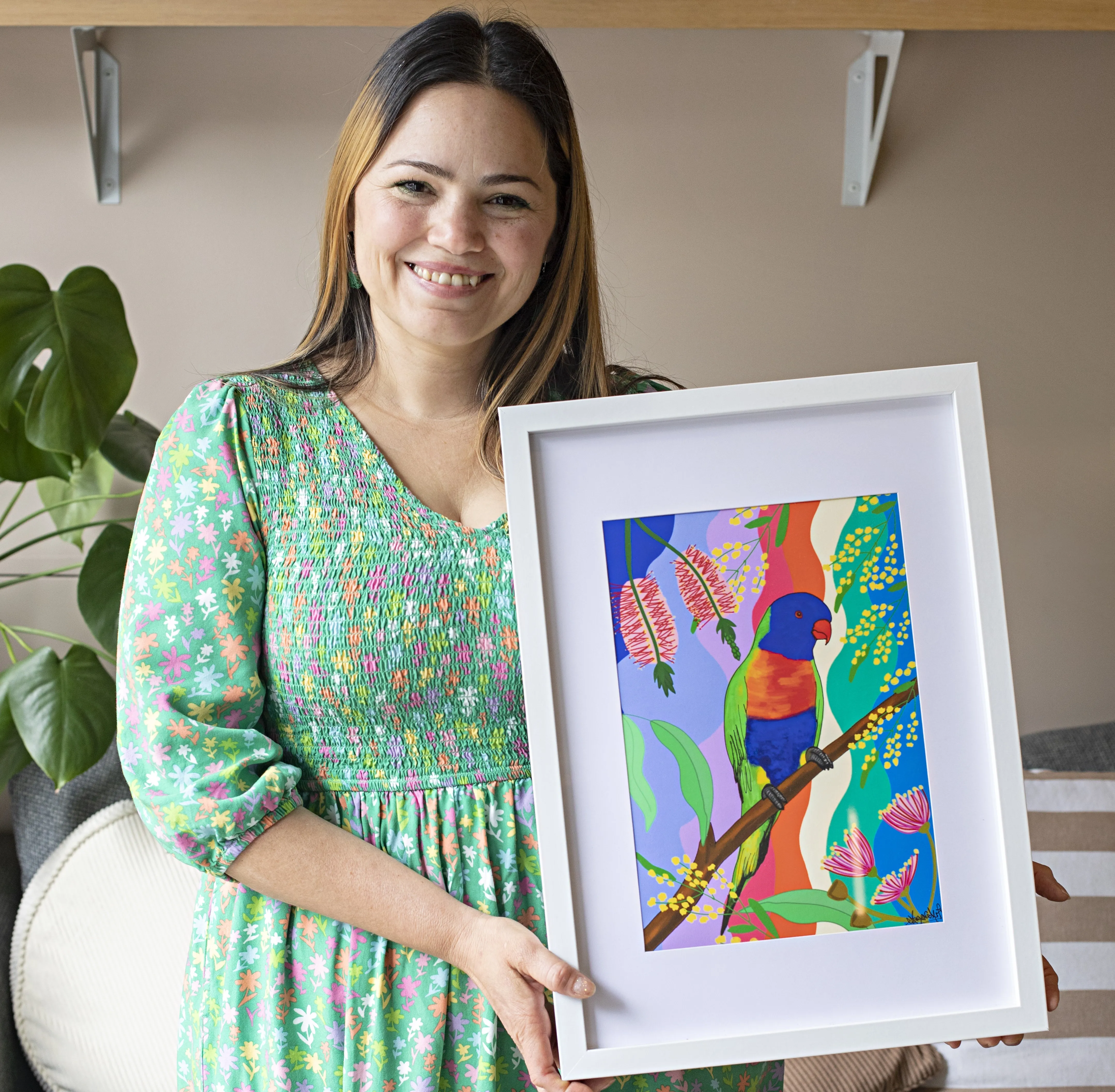 A woman in a green patterned dress holding a framed, colourful painting of a bird surrounded by bright botanical patterns.