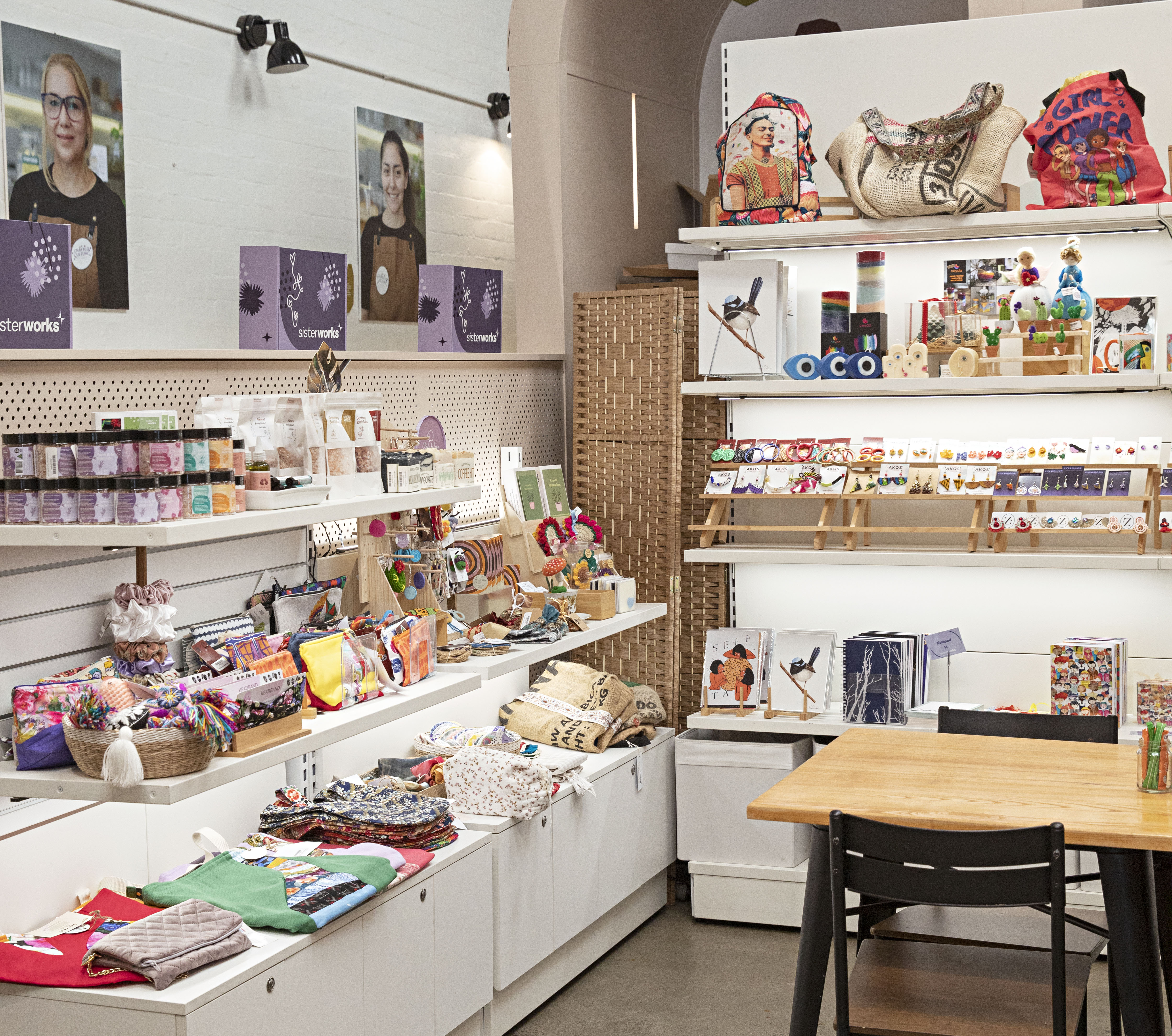 Shelves filled with handmade crafts, textiles, artwork and gift items inside the SisterWorks retail space, with a table and chair in the foreground.