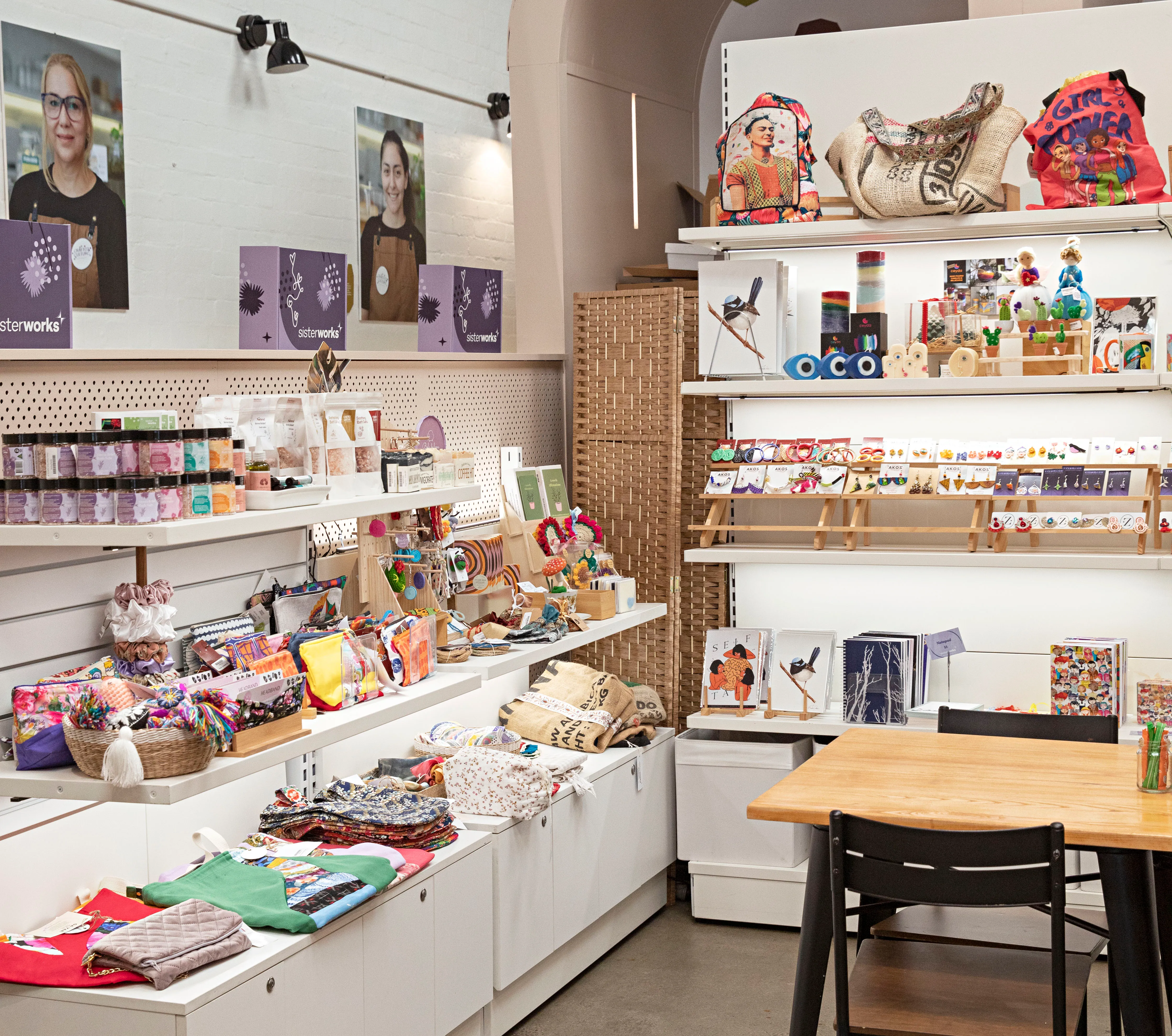 Shelves filled with handmade crafts, textiles, artwork and gift items inside the SisterWorks retail space, with a table and chair in the foreground.