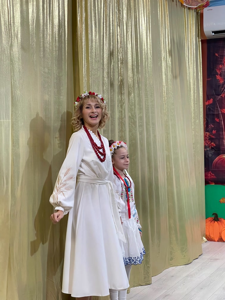 Eine Schülerin der Schule „Kinder der Zukunft“ im bestickten Hemd zusammen mit ihrer Lehrerin vor goldenen Kulissen in den Schulferien