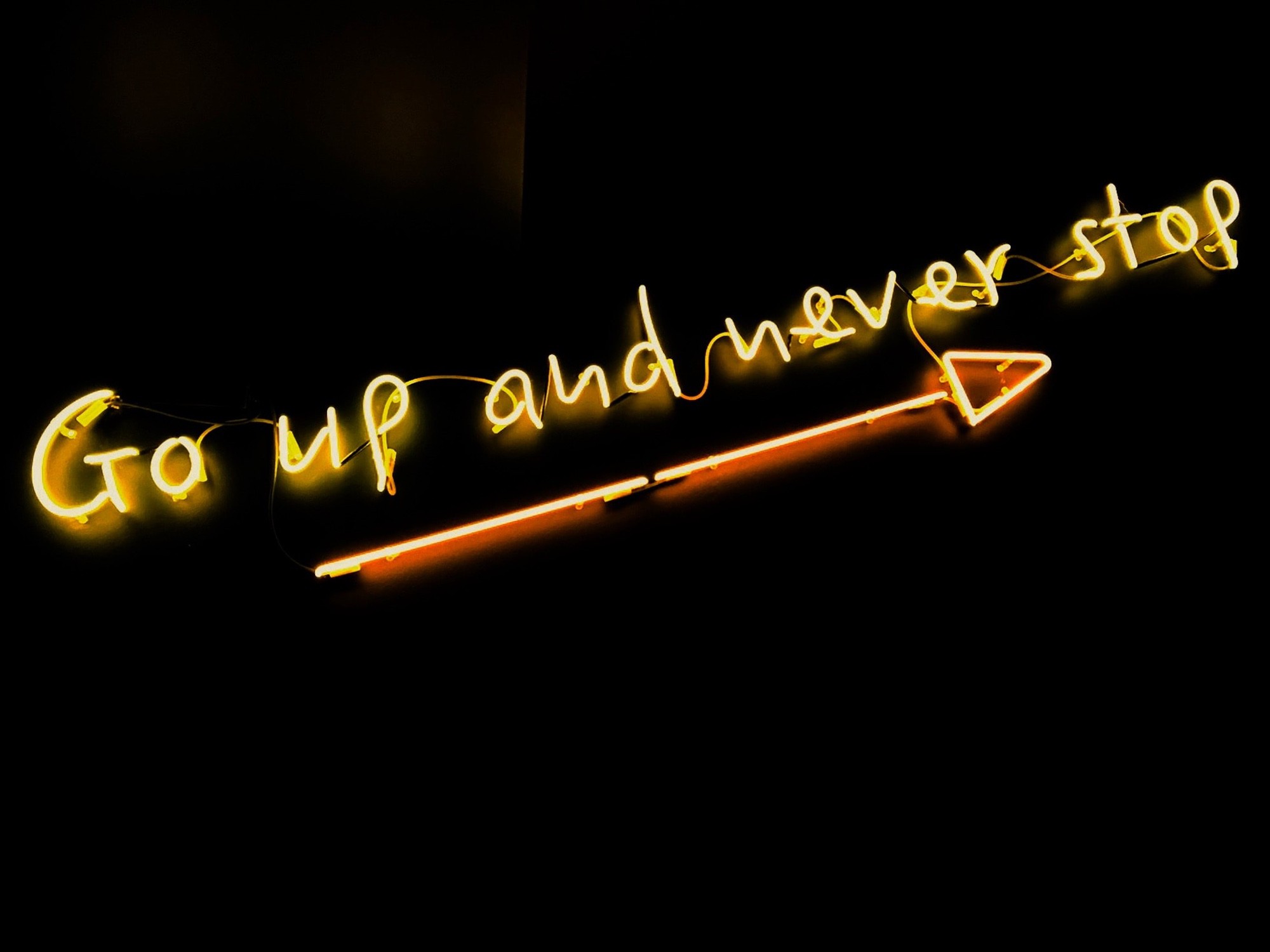 Neon sign saying 'Go up and never stop' against black background