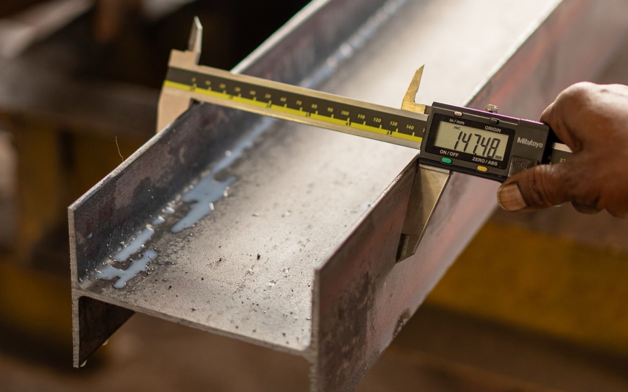 Digital caliper measuring the thickness of a structural steel I-beam in a fabrication workshop