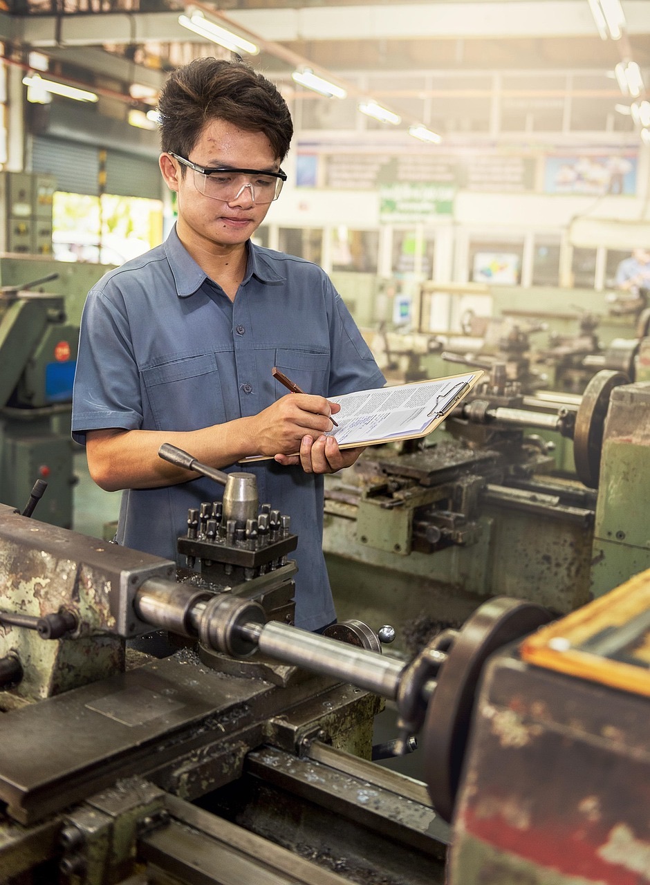 Quality engineer recording inspection results on a clipboard beside a lathe in a metal fabrication workshop