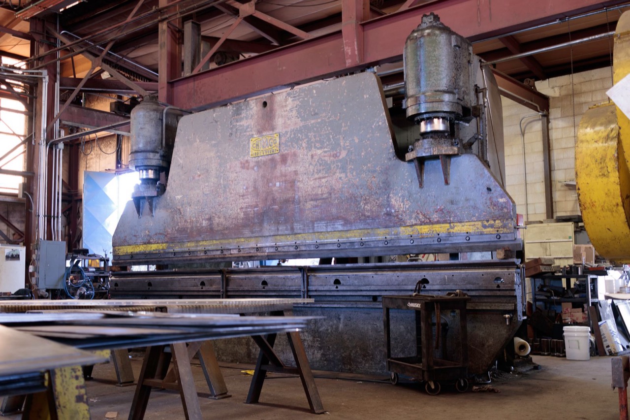 Large industrial press brake in a fabrication workshop with sheet metal stacked on a table in the foreground