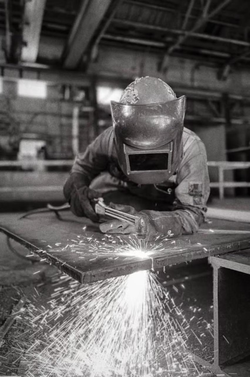 Fabricator welding a sheet metal panel on a workbench with sparks scattering across the shop floor