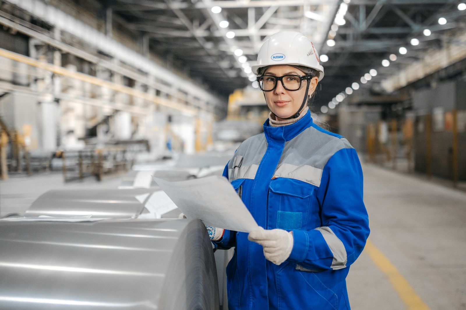 Quality engineer in a hard hat reviewing an inspection document beside rolled steel coils in a steel plant