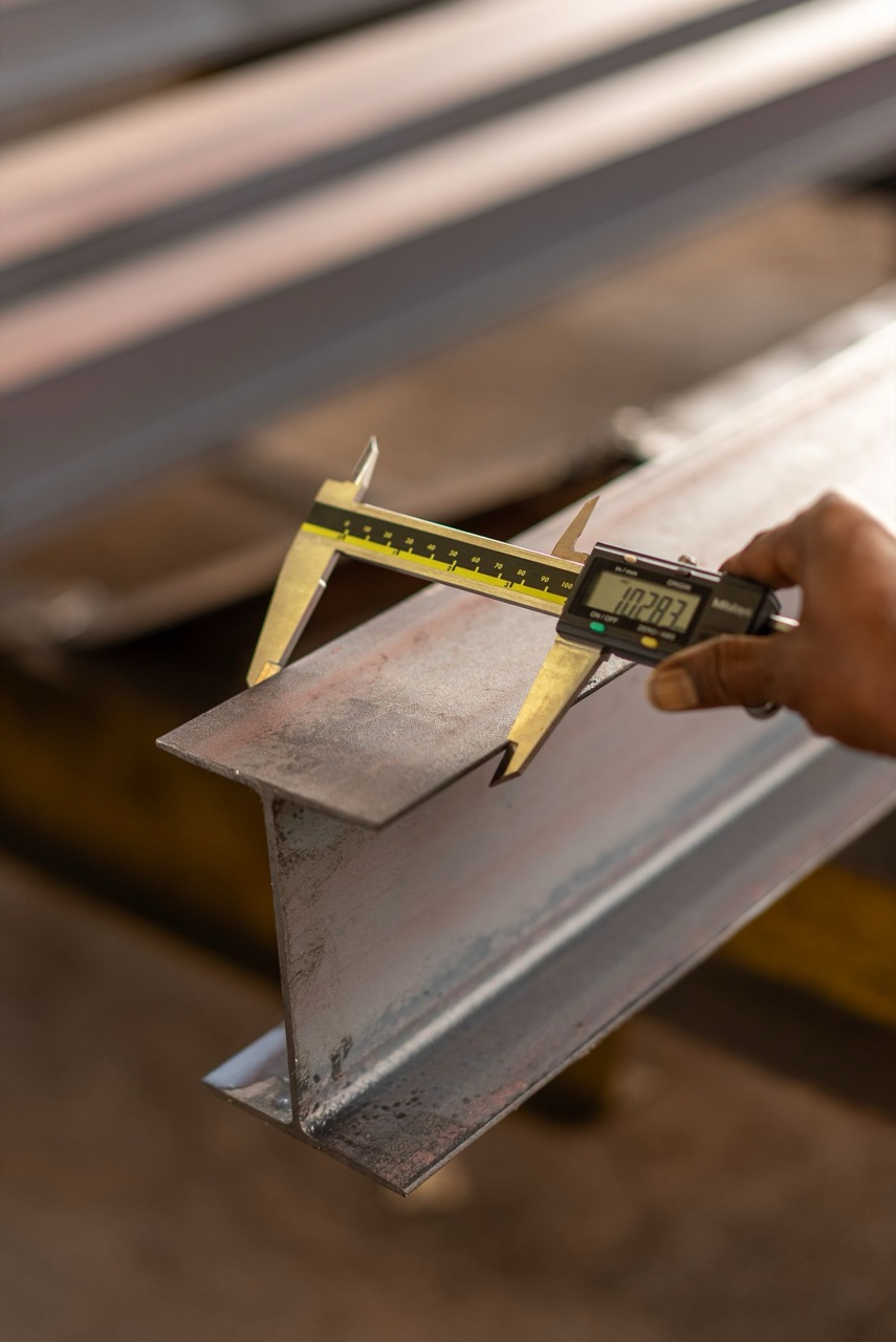 Digital caliper measuring the flange thickness of a steel I-beam on a fabrication shop floor