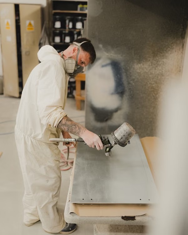 Technician in protective gear applying a spray finish to a metal panel