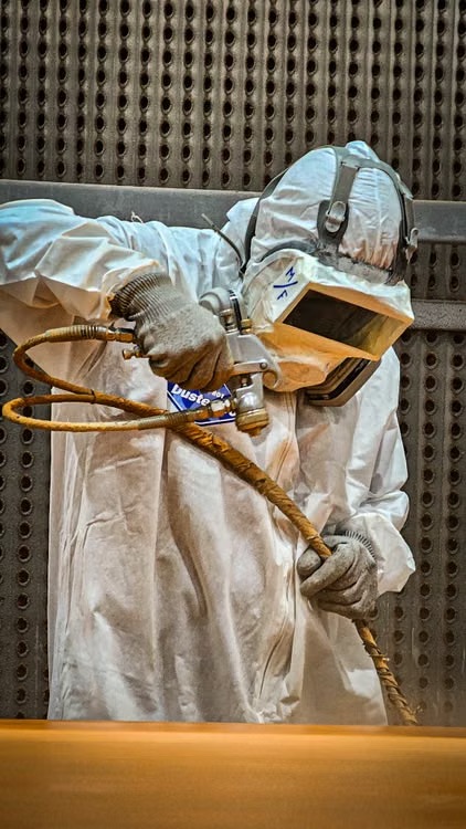 Worker wearing protective equipment sandblasting a metal surface in an industrial finishing booth