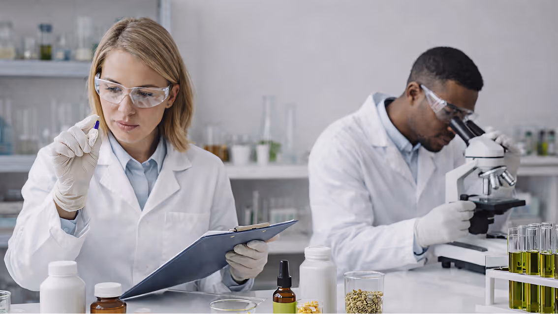 Two scientists in lab coats and safety glasses working in a laboratory; one examines a small vial while holding a clipboard, the other looks through a microscope amid various bottles and test tubes.