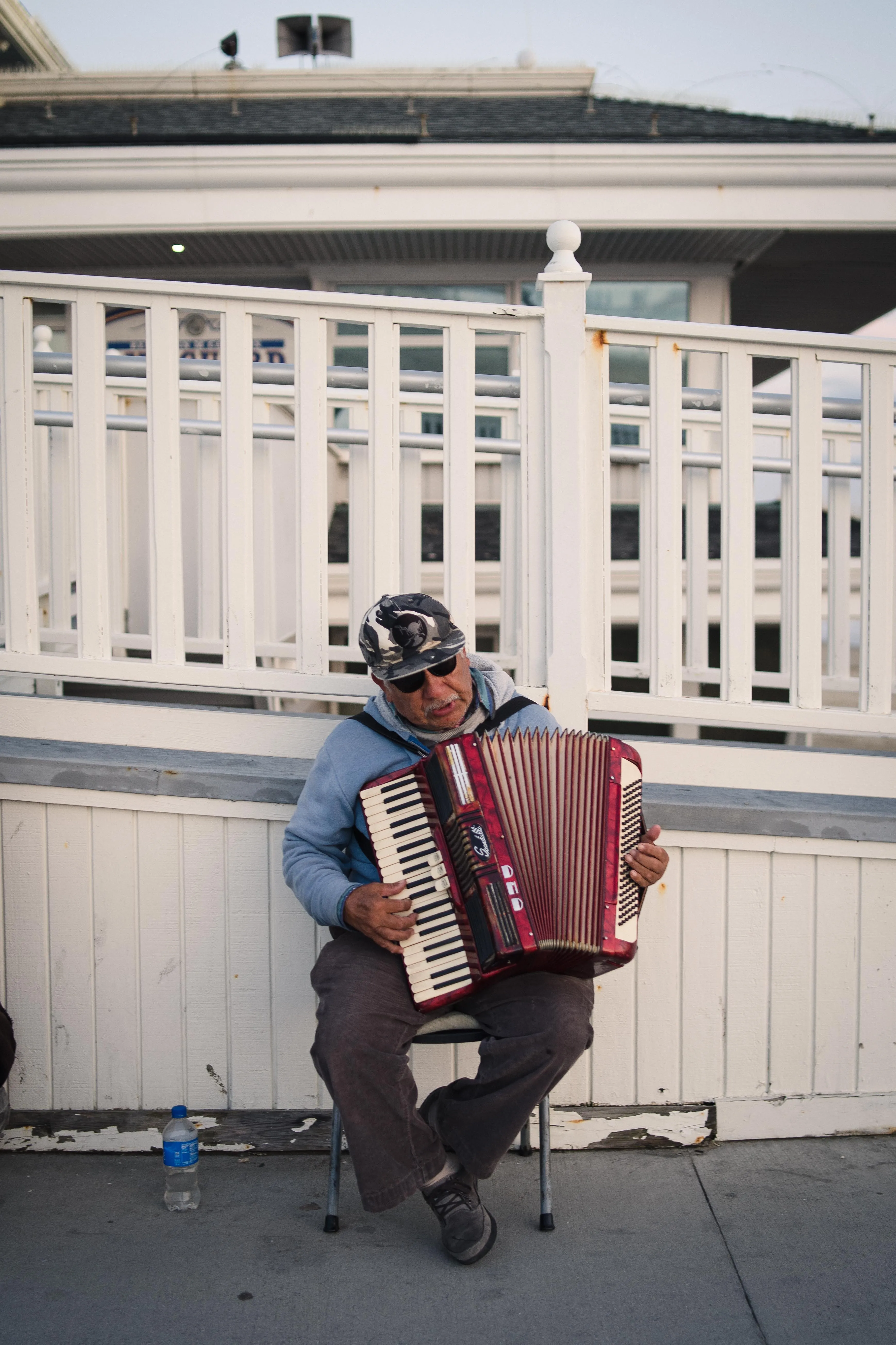 A street musician sitting on a chair playing an accordion beside a white wooden railing.