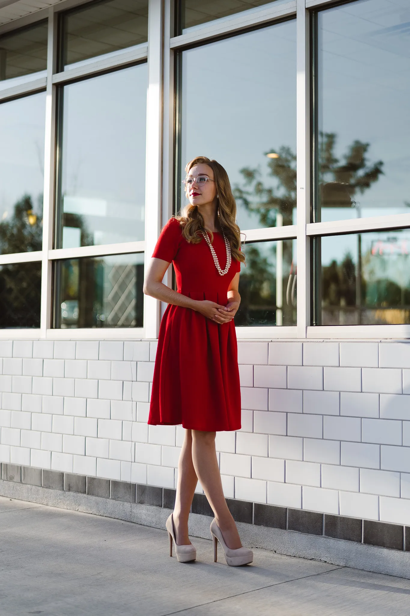 A woman in a red dress standing outside a building with large reflective windows, gazing off into the distance.