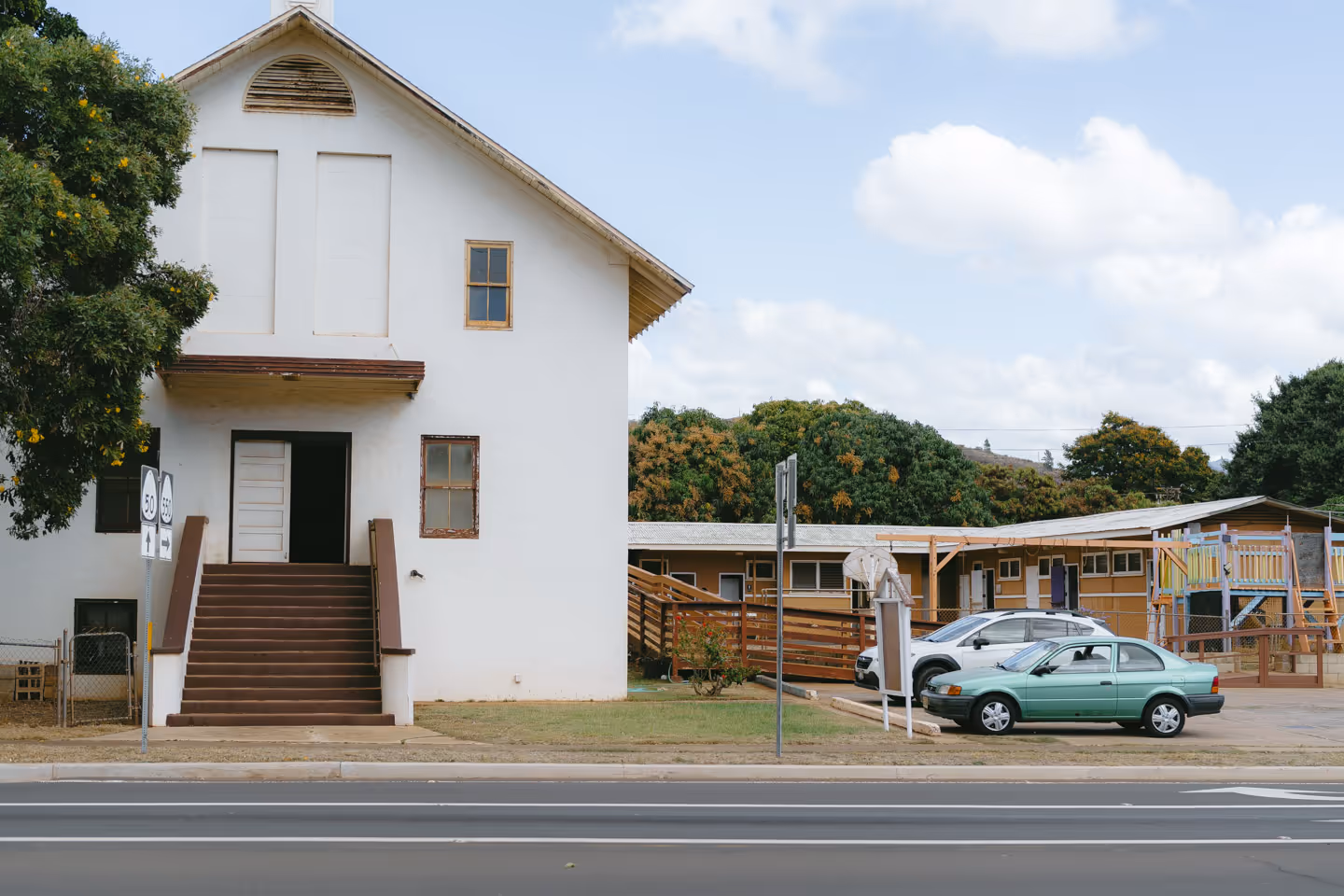 A white two-story building with brown steps and a parked car on a sunny day.