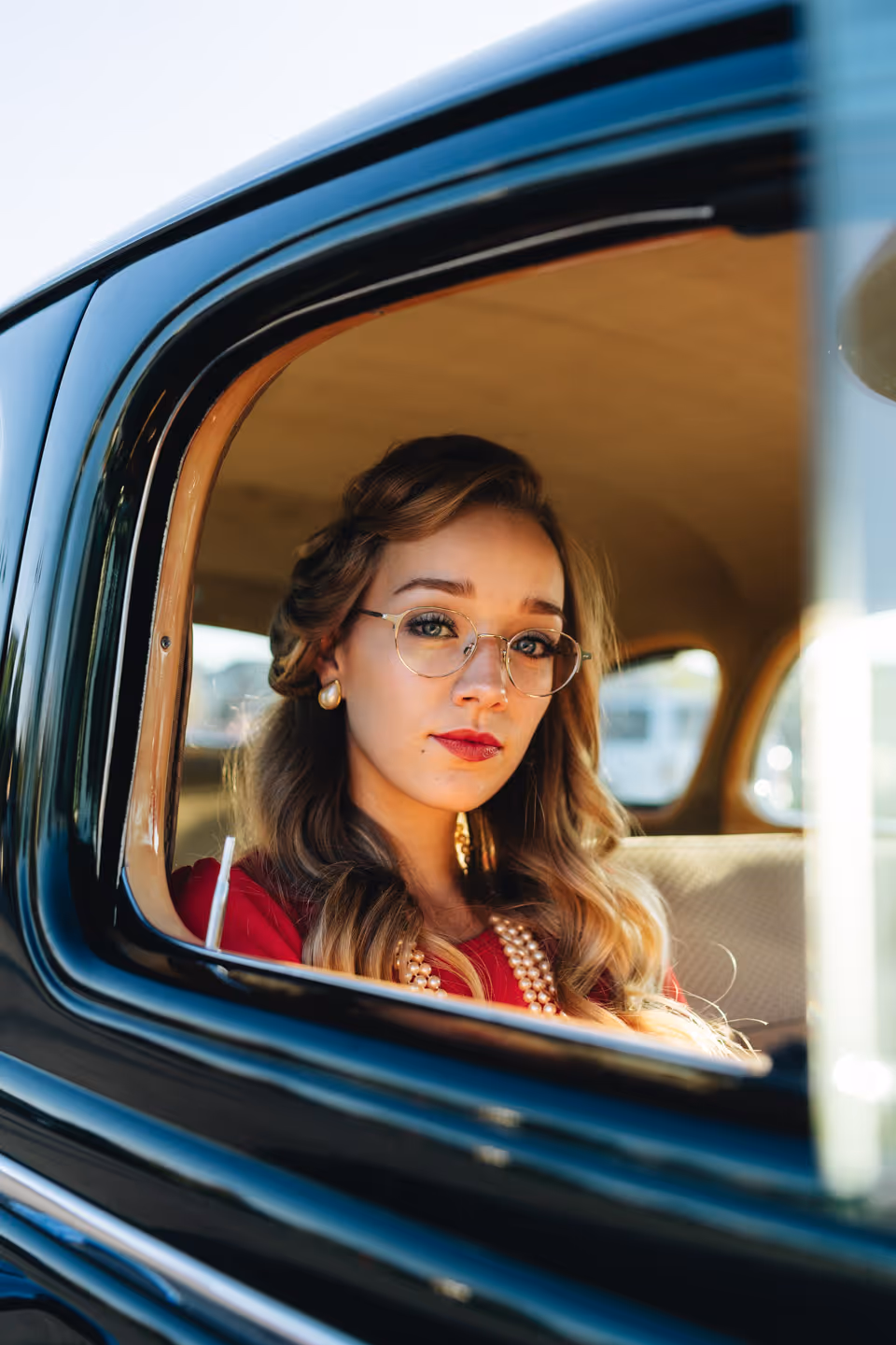 A woman in a red dress and glasses looking out the window of a vintage black car, sunlight reflecting off the glass.