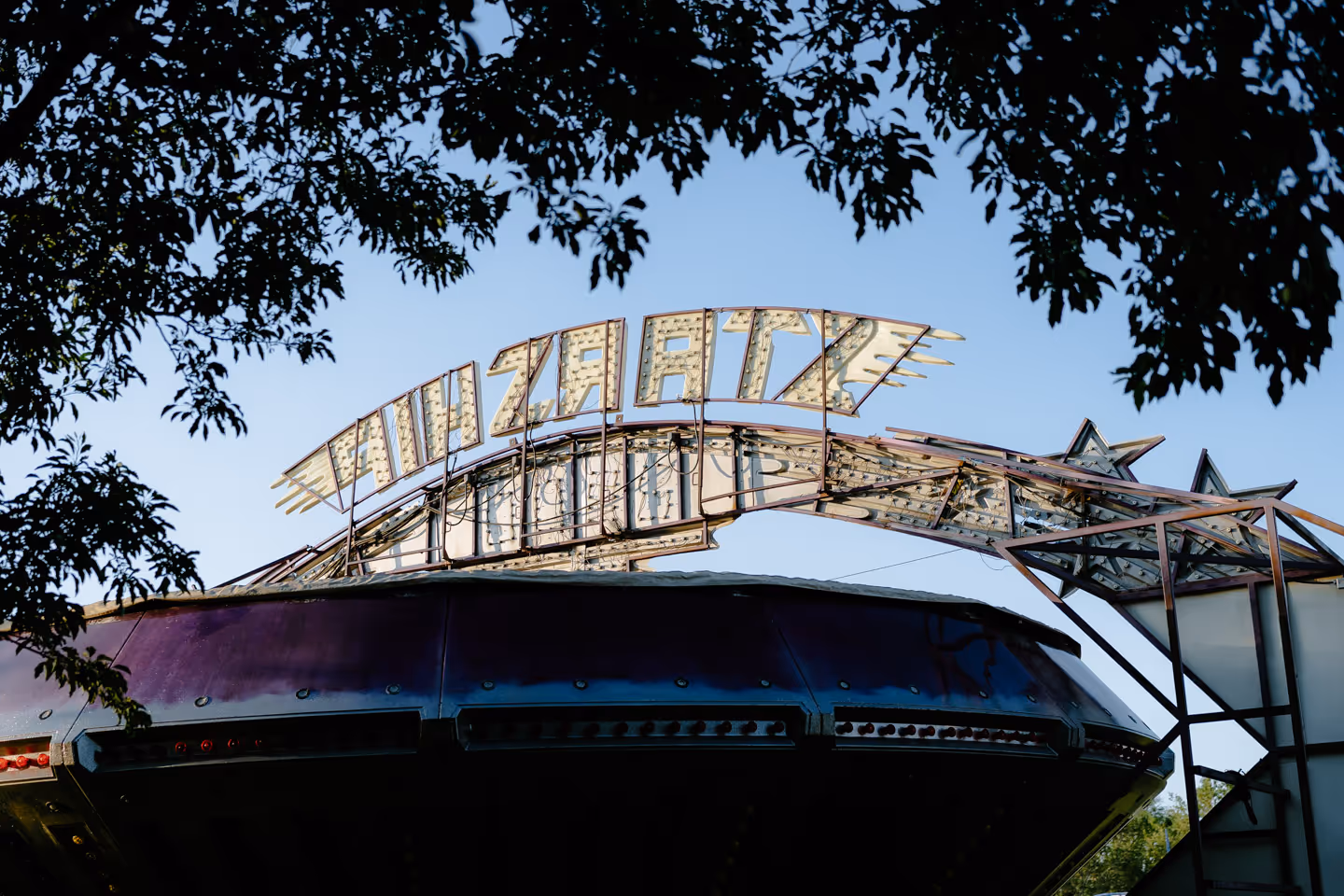 A close-up of an old carnival ride with a faded sign reading “Tilt-A-Whirl,” partially framed by tree branches.