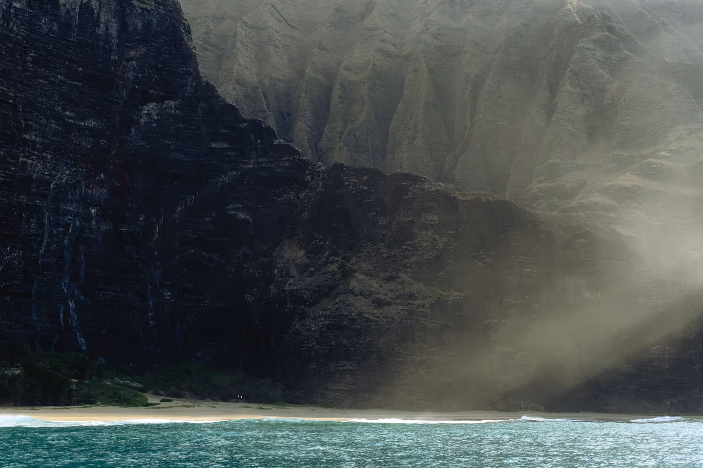 Steep rocky coastal cliffs with sunlight streaming through mist above turquoise water.