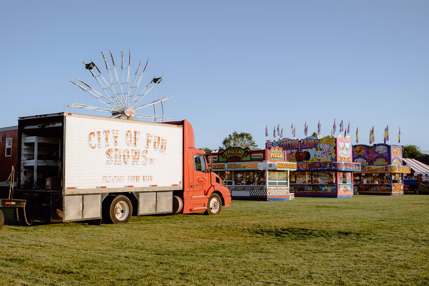 A red truck with a sign reading “City of Fun Shows” parked on grass beside colorful carnival booths in the distance.